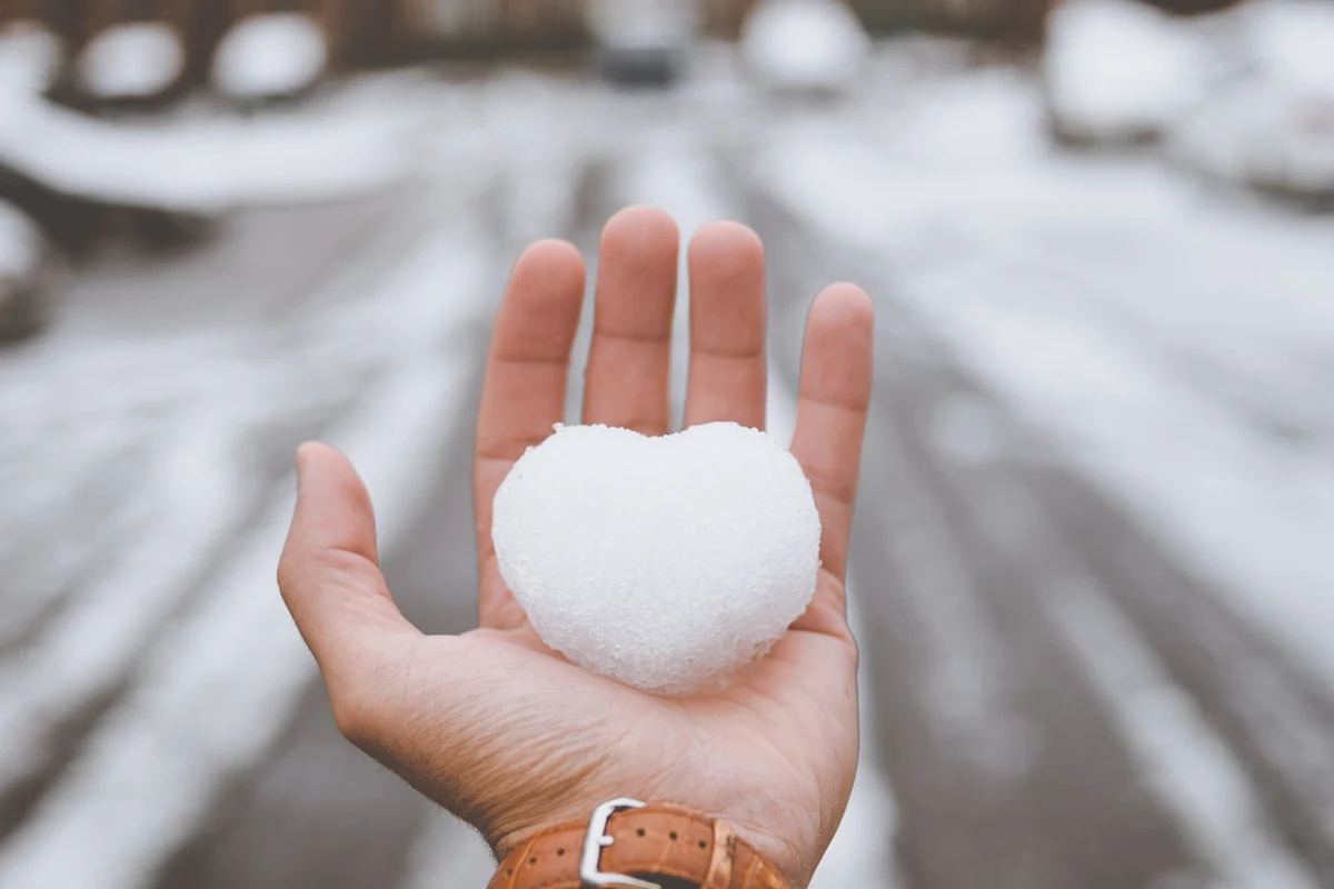 An open hand holding a heart-shaped ball of snow on a snowy street